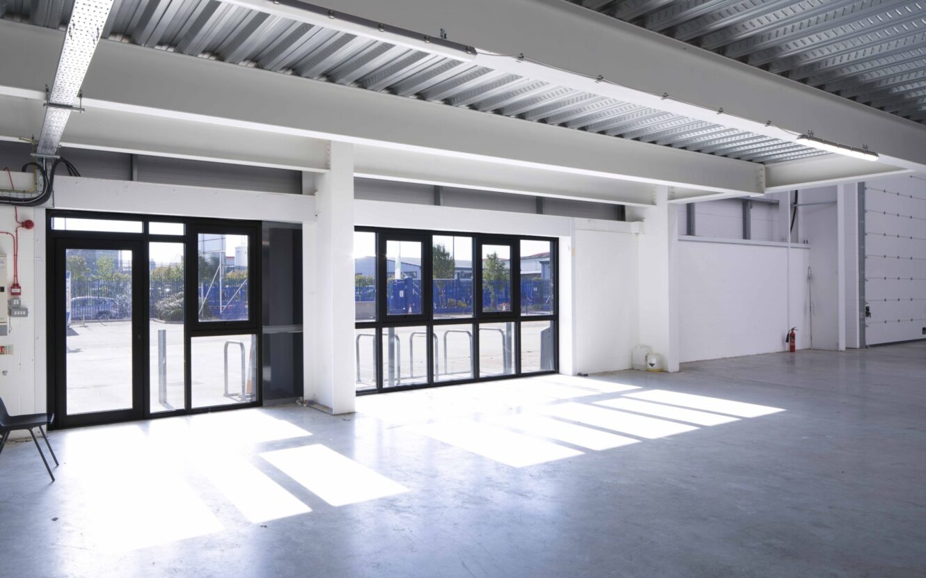 Large, empty industrial space with polished concrete floors, high ceilings, and large windows letting in natural light. Bike racks and trees are visible outside.