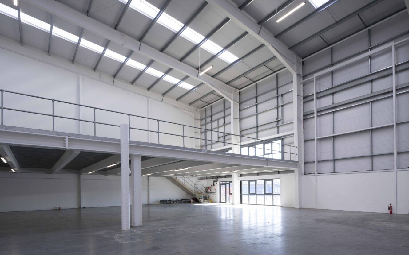 Interior of a large, empty industrial warehouse with high ceilings, upper mezzanine level, metal railings, and large windows letting in natural light.