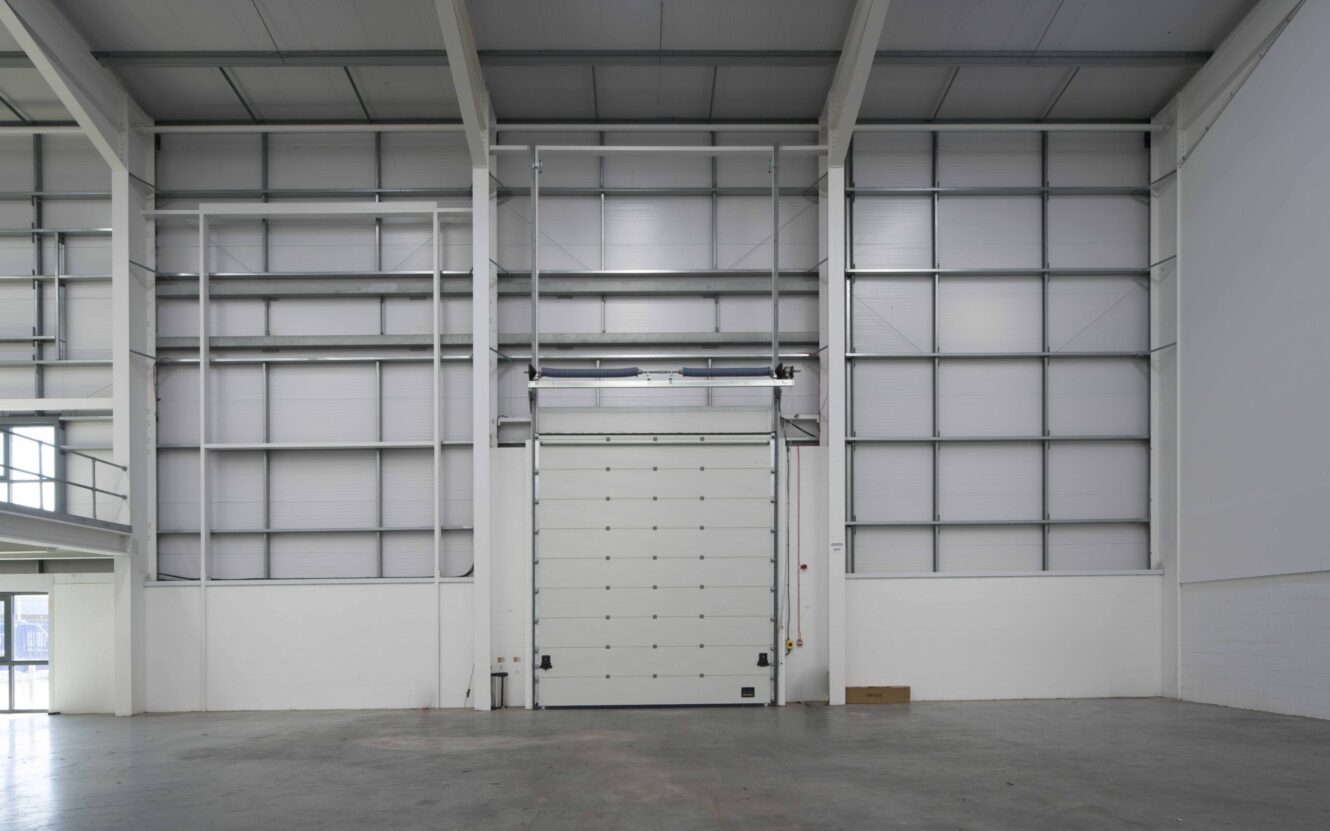 Interior view of an empty industrial warehouse with a large closed loading bay door, high ceiling, and metal wall panels.