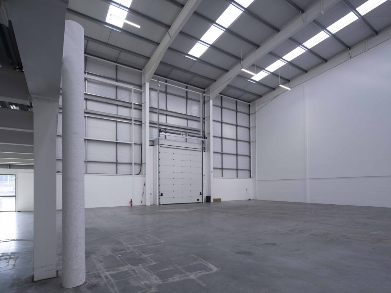 Interior of an empty industrial warehouse with high ceilings, skylights, concrete floor, and a large roller shutter door.