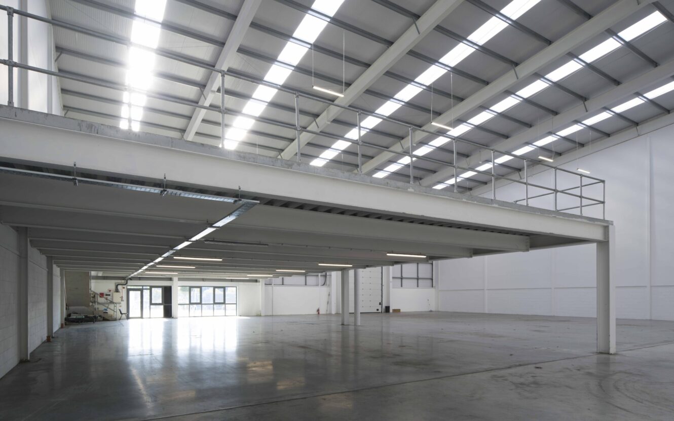 Spacious, empty industrial warehouse interior with high ceilings, metal beams, mezzanine level, and large windows letting in natural light.