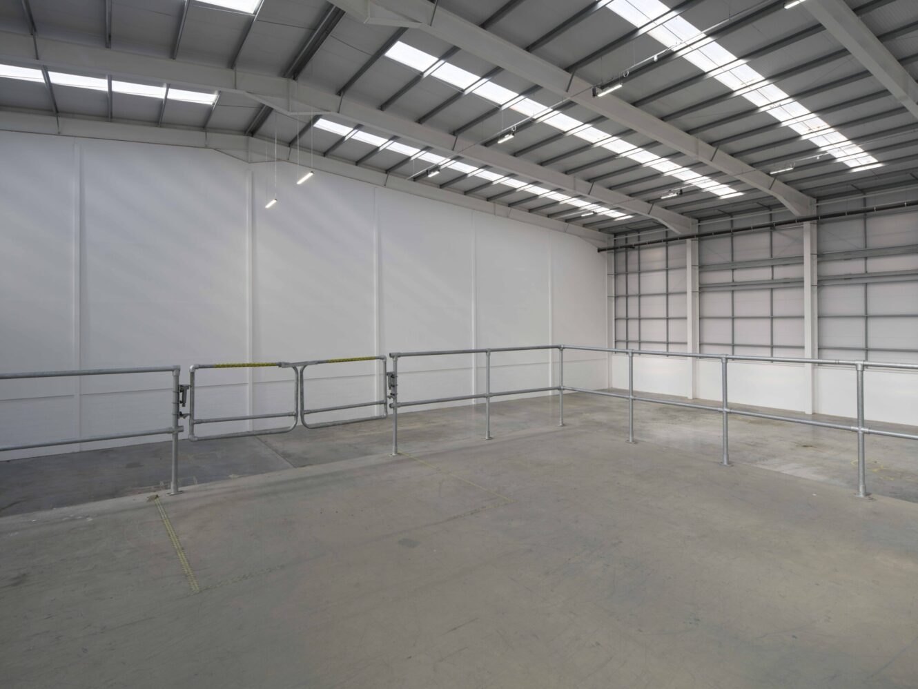 An empty industrial warehouse interior with concrete flooring, metal railings, and a high ceiling with skylights.