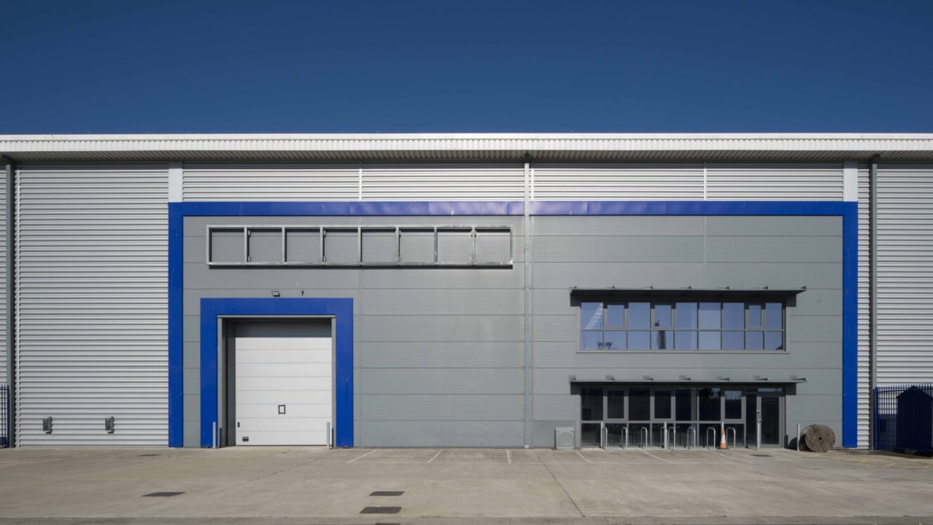 Front view of a modern industrial warehouse with a large blue-framed loading bay door, windows, and an empty concrete forecourt under a clear blue sky.