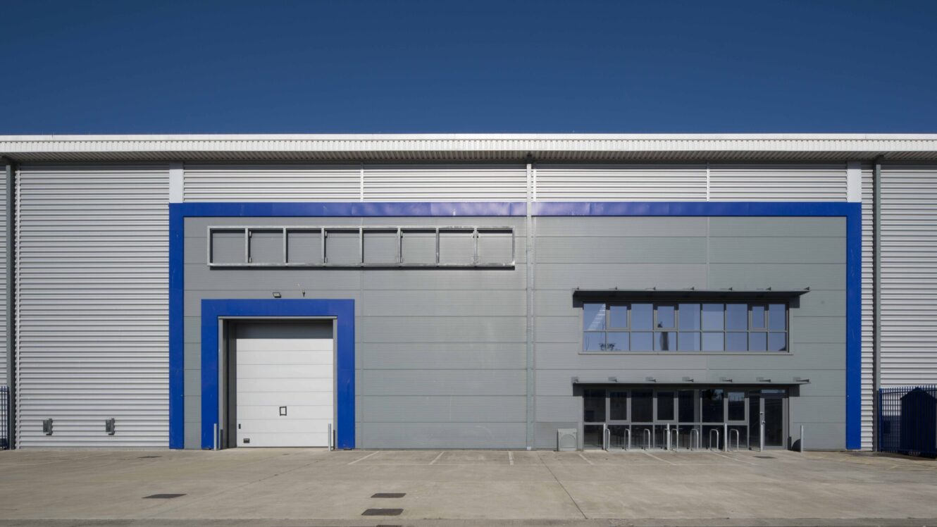A modern industrial warehouse with a large white loading bay door, grey metal cladding, blue trim, and multiple windows under a clear blue sky.