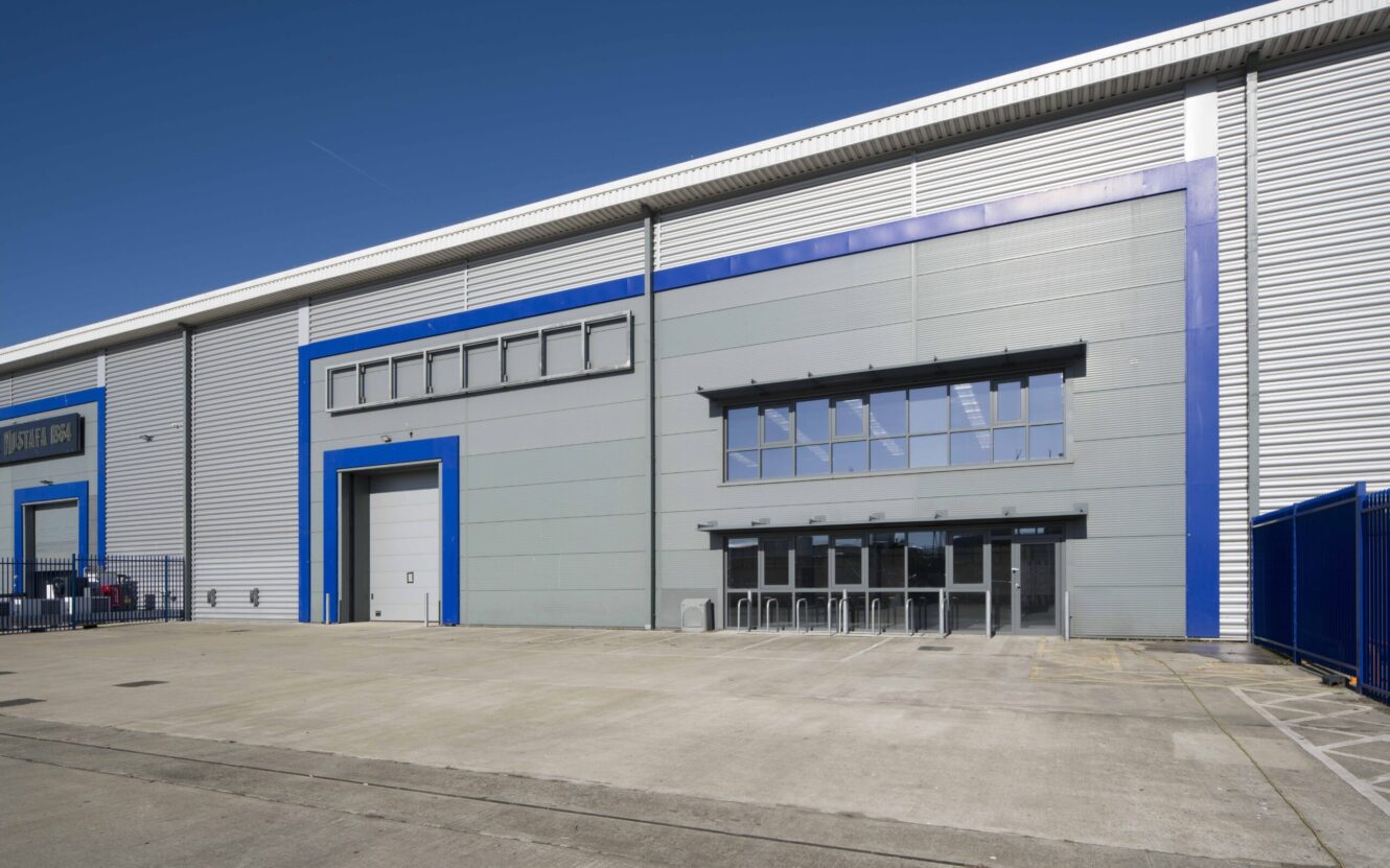 Large industrial warehouse exterior with grey walls, blue trim, a loading bay door, multiple glass entrance doors, and a wide concrete area in front.
