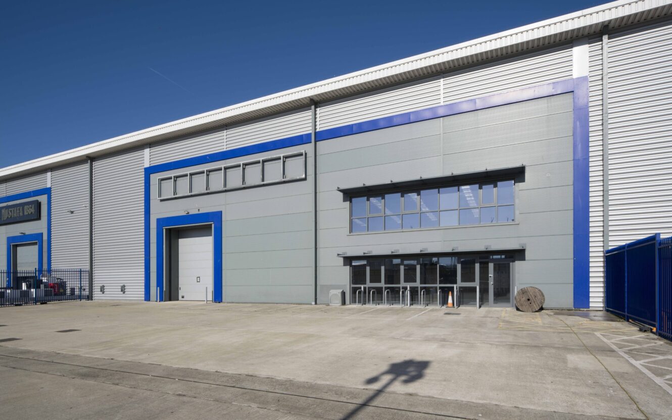 Large industrial warehouse building with grey walls, blue trim, a loading bay door, glass-fronted entrance, and a wide empty concrete forecourt in front under clear blue sky.