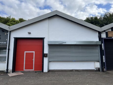 A white industrial building with a red door and closed silver shutter, set against a cloudy sky with trees in the background.