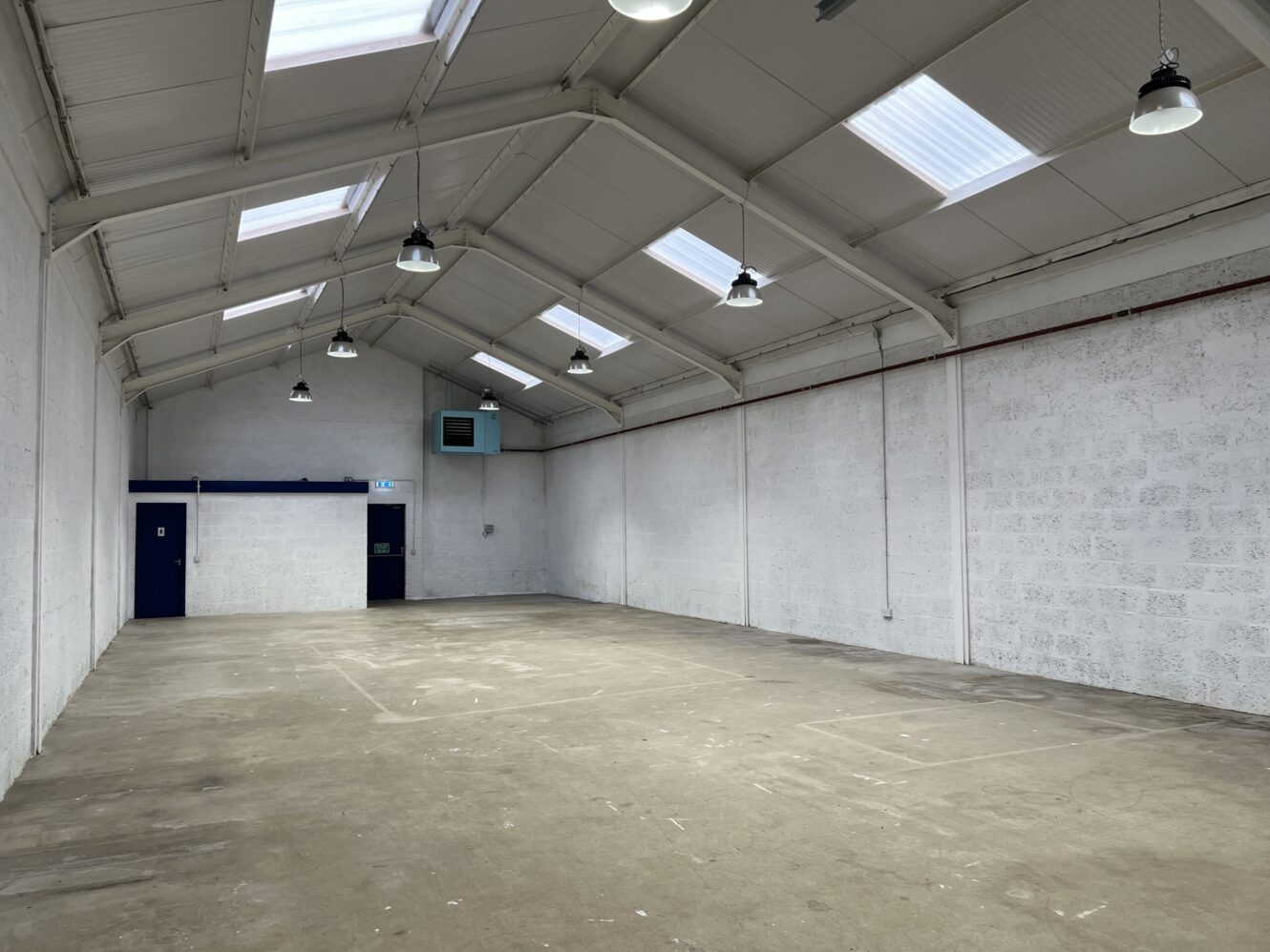Empty industrial warehouse with white walls, concrete floor, high ceiling, skylights, hanging lights, and a blue door at the far end.