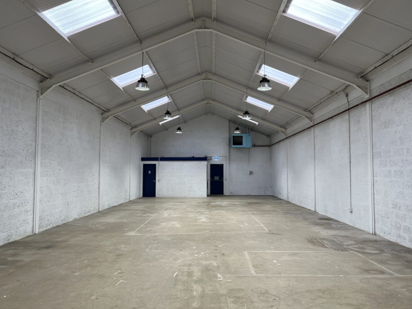 Empty warehouse interior with white walls, high ceilings, skylights, exposed beams, and overhead lighting. A blue door and fire exit are visible at the far end.