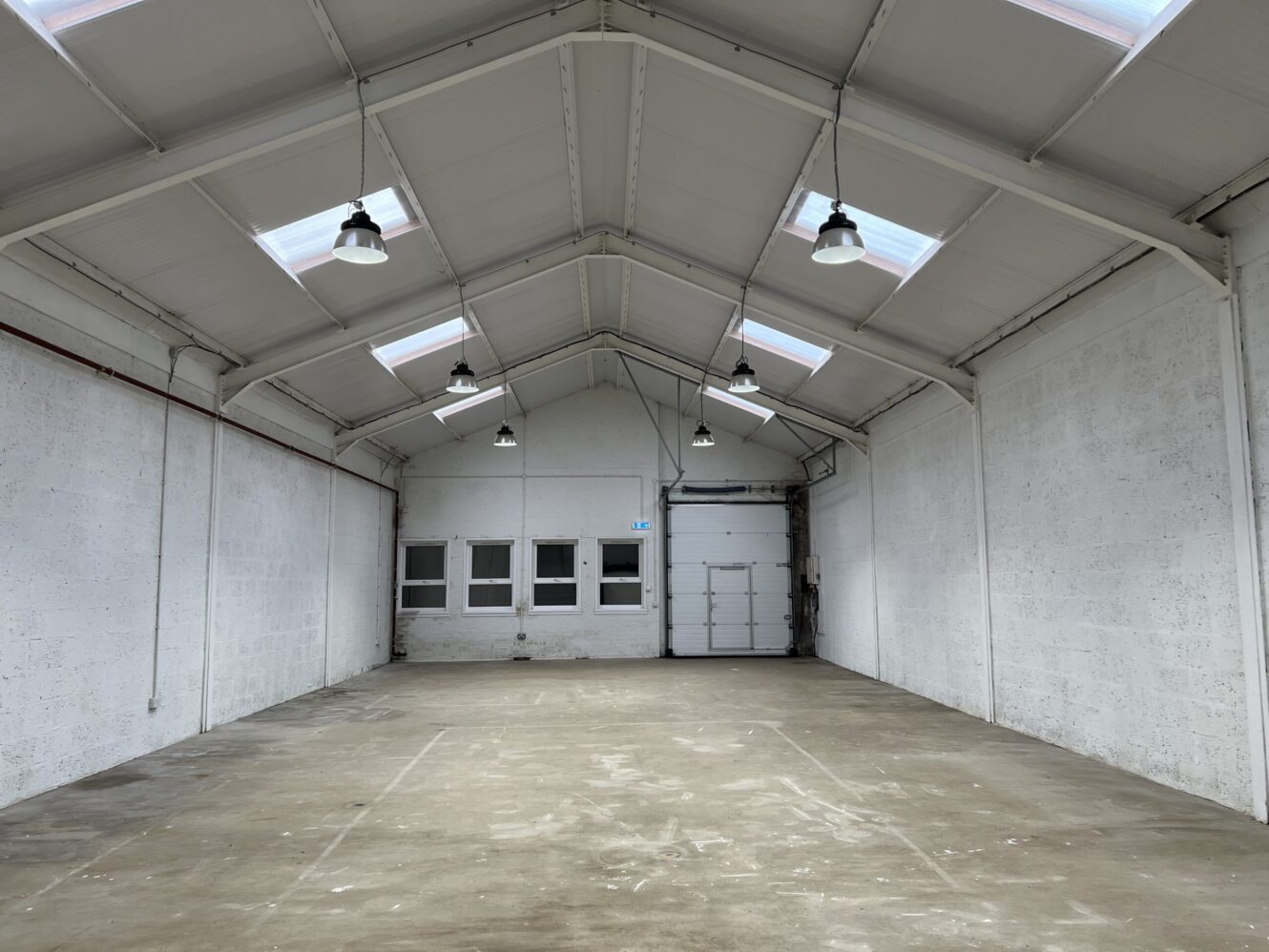 Interior of an empty industrial warehouse with white walls, high ceiling, overhead lights, rooflights, and a closed garage door at the far end.