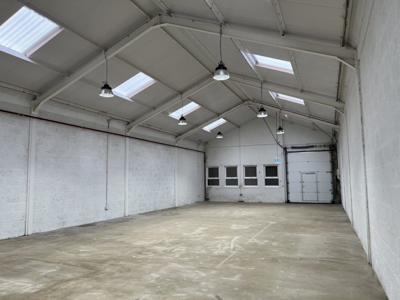 Empty industrial warehouse with white walls, high ceiling, skylights, concrete floor, ceiling lights, and a garage door at the far end.