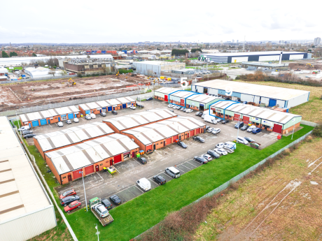 Aerial view of an industrial estate with multiple warehouse units, parked cars in front, and construction activity visible in the background.