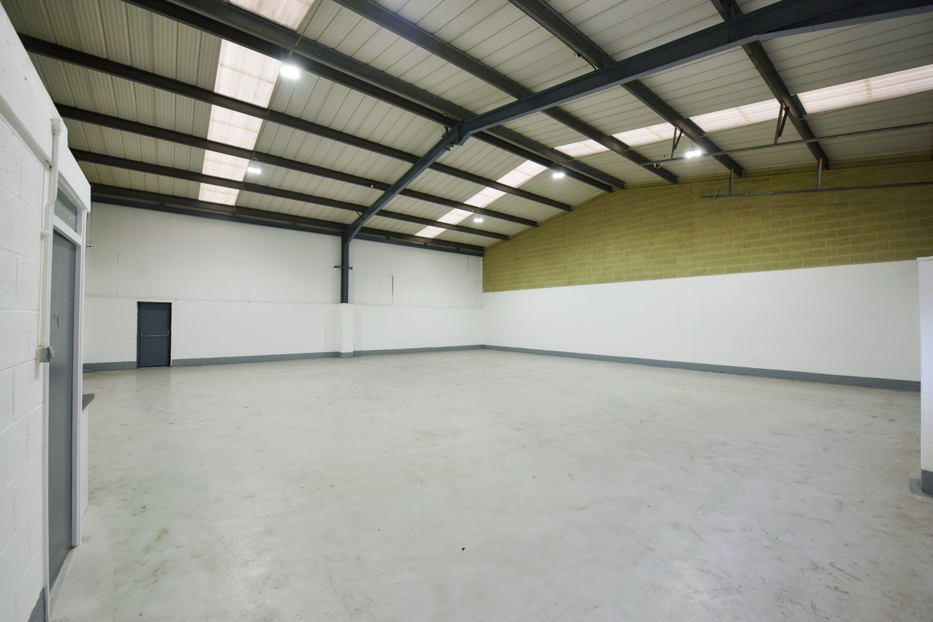 Empty industrial warehouse with white walls, high ceiling, exposed beams, and concrete floor. Fluorescent lights are mounted on the ceiling, and there is a small door on the left wall.