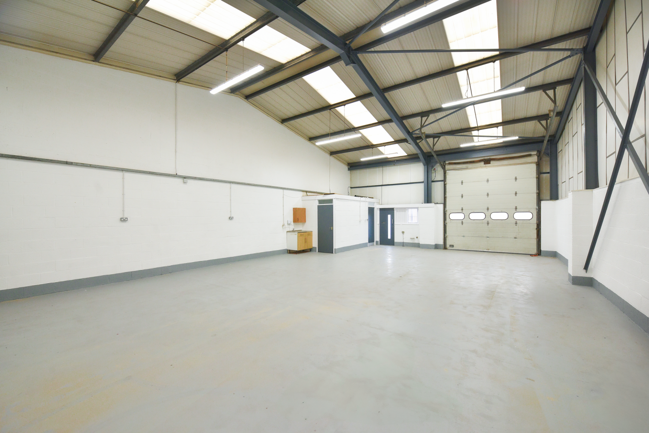 Empty industrial warehouse with white walls, high ceilings, large windows, overhead lights, a small kitchenette area, and a closed roller shutter at the far end.