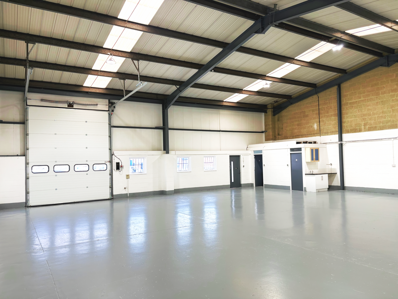 Interior of an empty industrial warehouse with a large roller shutter door, high ceiling, small office area, and polished concrete floor.