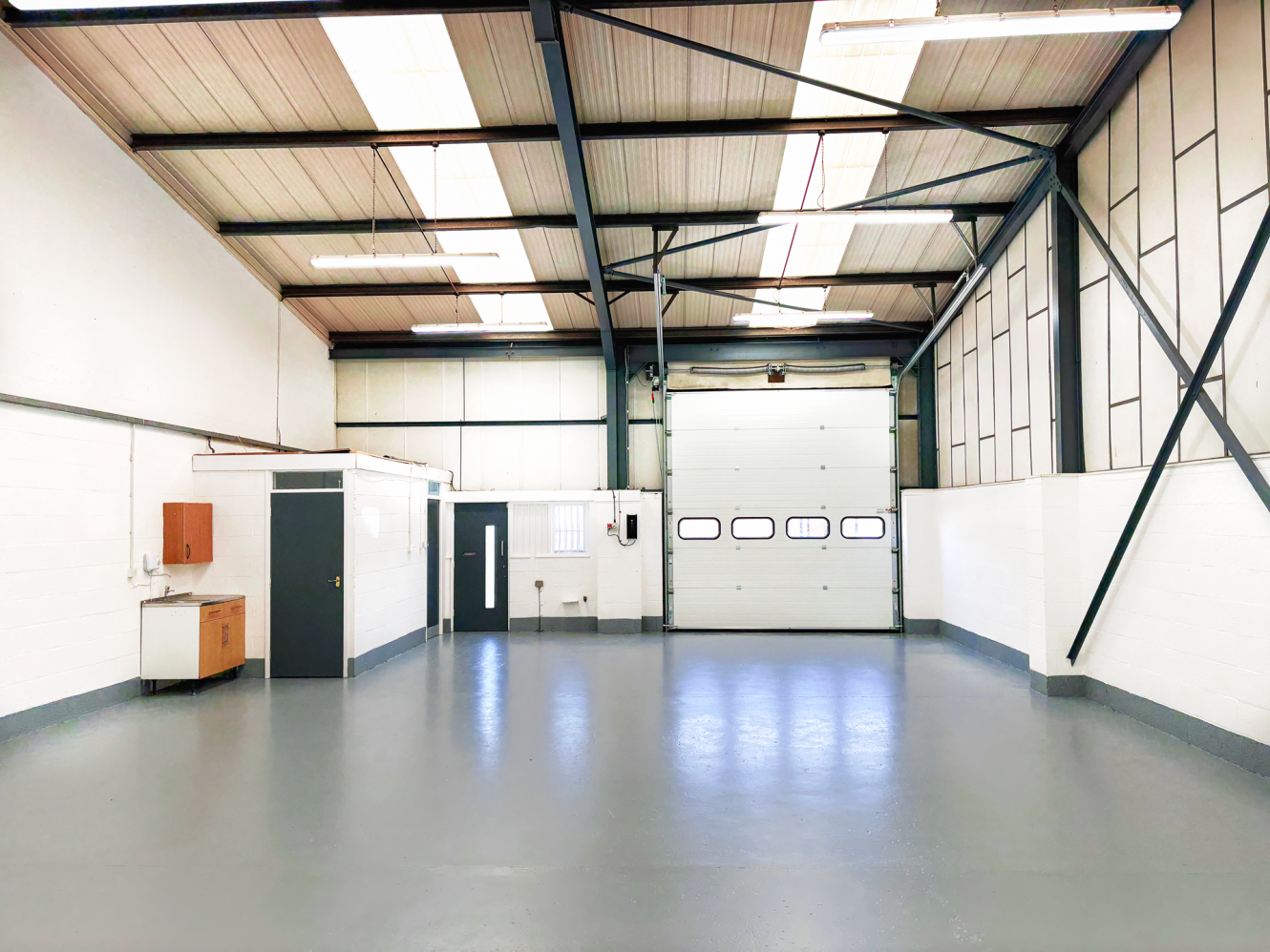 Empty industrial warehouse unit with white walls, grey floor, high ceiling, large roller shutter door, and small kitchenette area on the left. Ceiling lights and windows provide ample illumination.