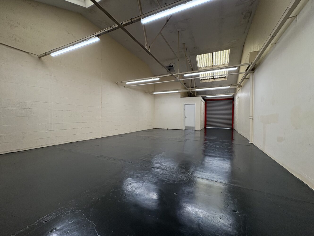 Empty industrial room with black glossy floor, beige walls, fluorescent ceiling lights, a white door, and a red roller garage door at the far end.