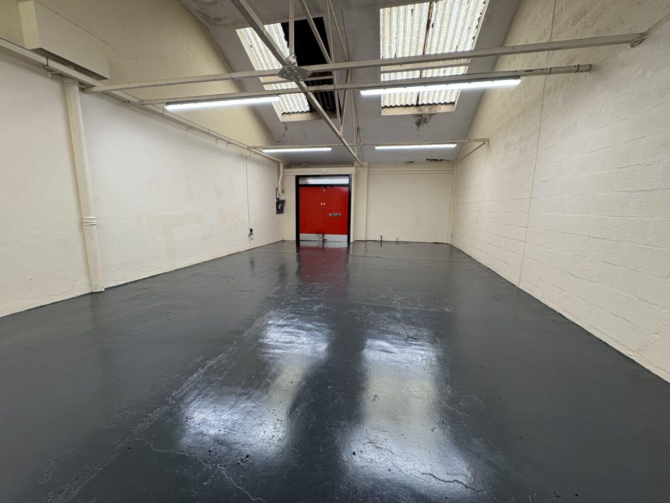 Empty warehouse room with glossy dark floor, white walls, exposed piping, overhead fluorescent lights, and a red double door at the far end.
