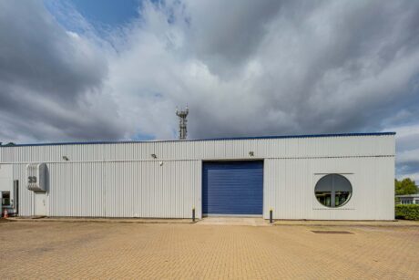 A white industrial warehouse with a blue roller shutter door, circular window, and cloudy sky above. Brick-paved area in front.