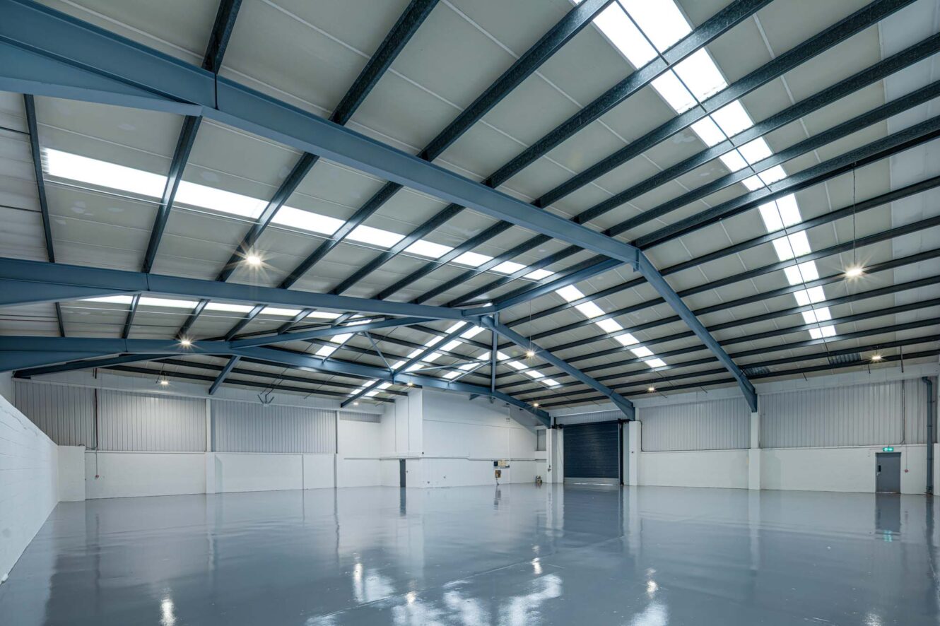 Interior of a large, empty industrial warehouse with high ceilings, exposed steel beams, skylights, and polished concrete floors.