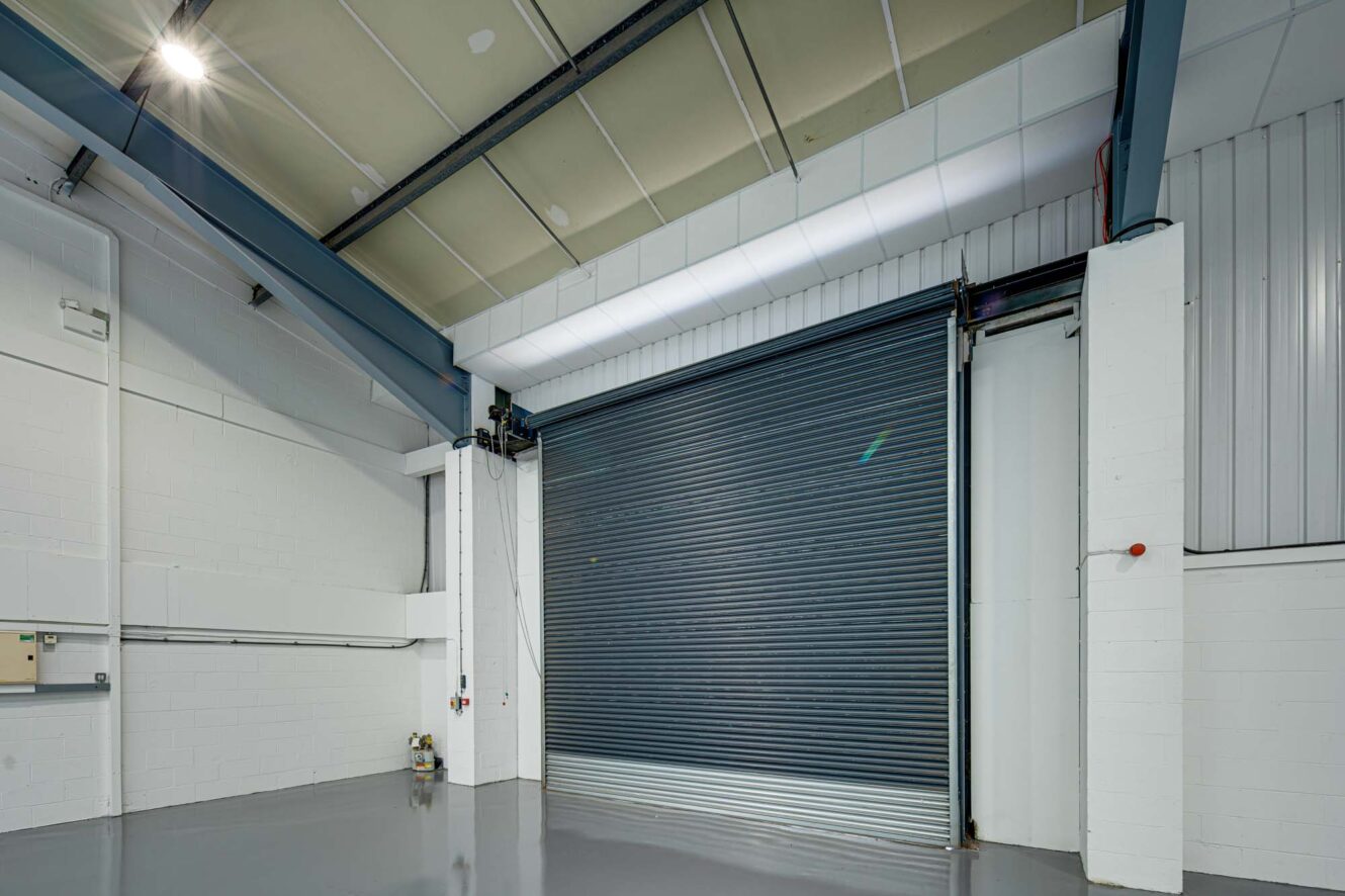 Interior of an industrial warehouse with a large closed metal roller shutter door, high ceiling, and clean, polished concrete floor.