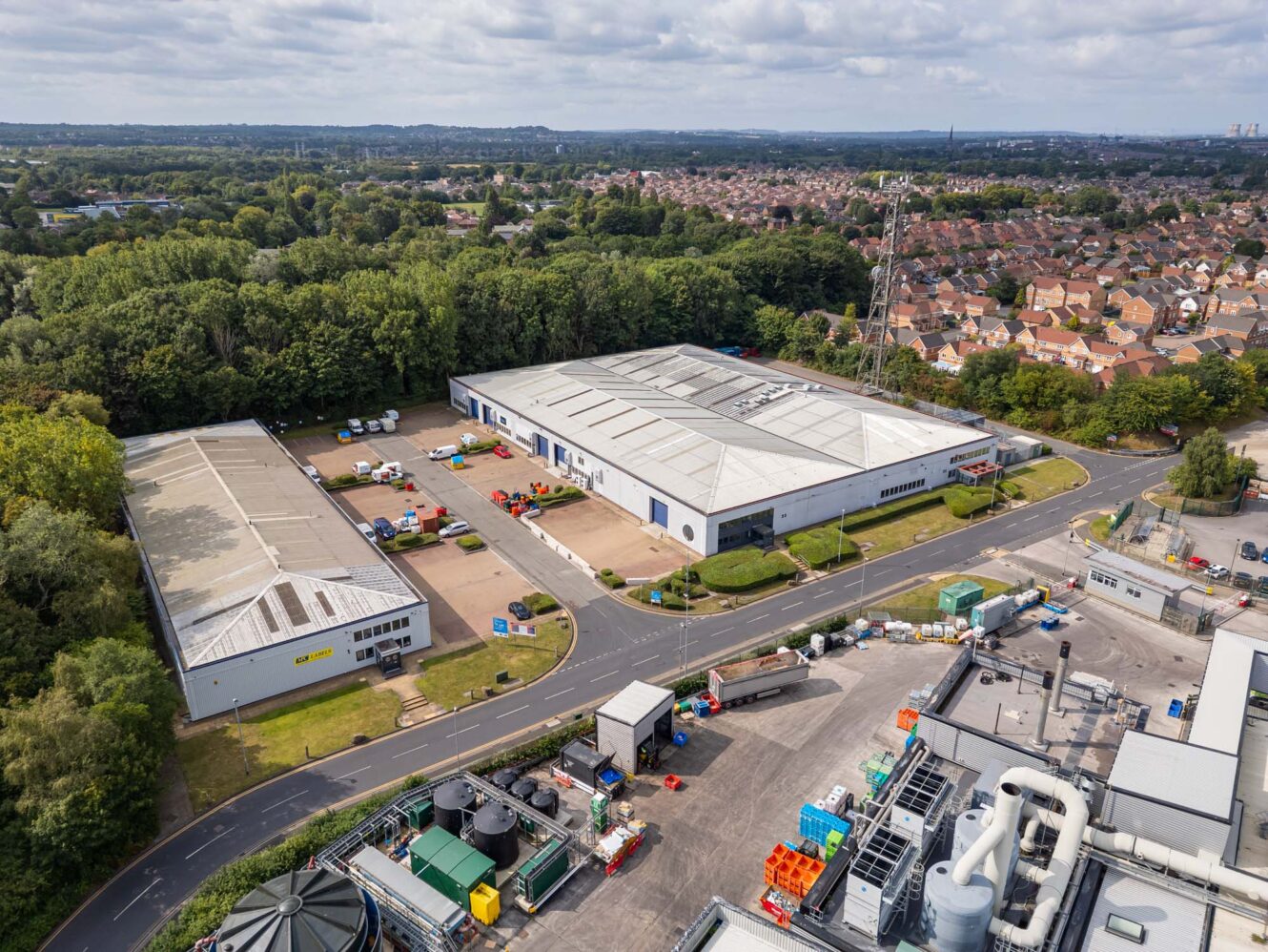 Aerial view of an industrial estate with warehouses, storage containers, and nearby residential houses bordered by trees under a cloudy sky.