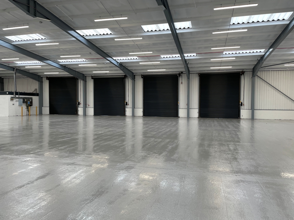 Large empty warehouse with a shiny concrete floor, white walls, overhead lights, and five closed black roller shutter doors along one side.