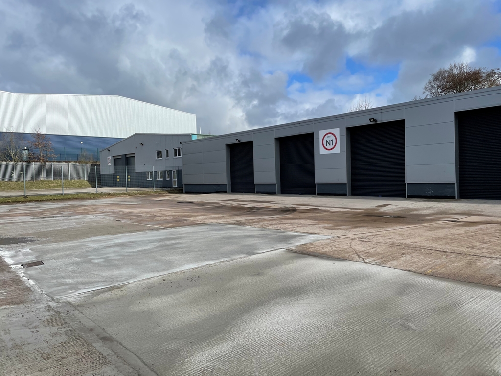 A modern industrial building with several large black garage doors, a No parking sign, and an empty paved car park under a partly cloudy sky.