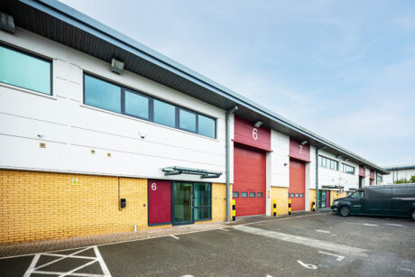 Modern industrial building with brick and white facade, large red roller shutters labelled 6, glass entrance, and parked black van in front.
