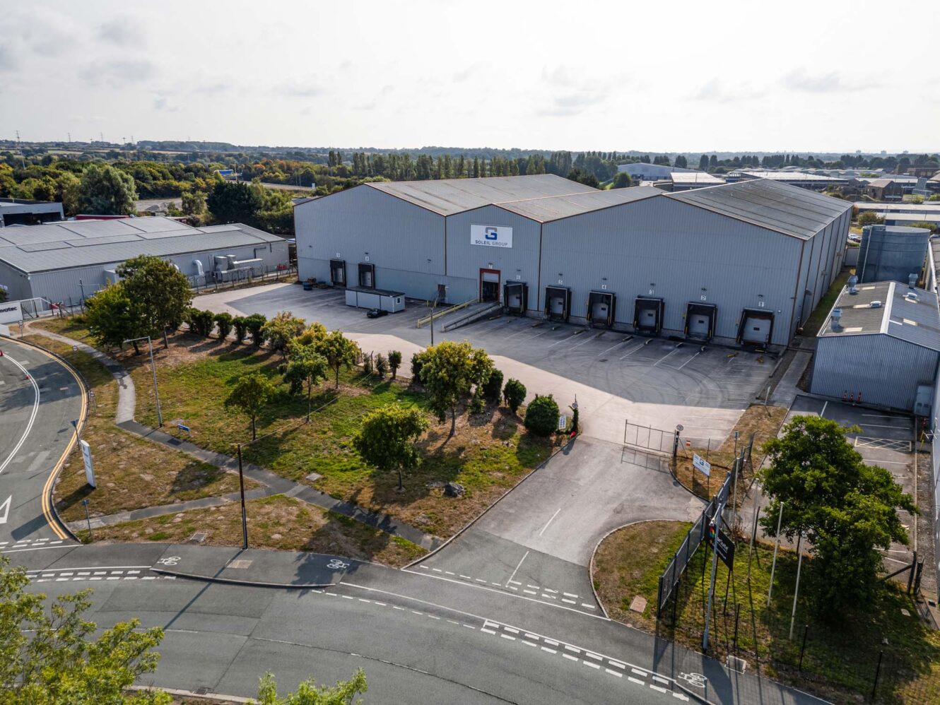 A large industrial warehouse with multiple loading bays and gated entrance, surrounded by trees and parking areas, situated near a curved road in a business park.
