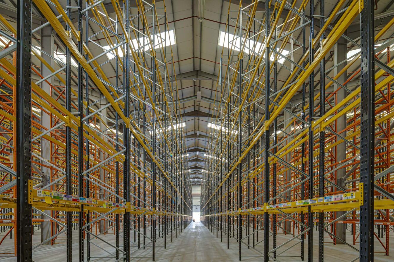 Interior view of an empty warehouse aisle with tall, metal storage racks on both sides under a high, well-lit ceiling.