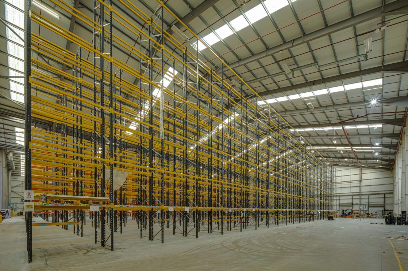 Large, empty warehouse interior with tall, yellow metal shelving racks and a high, industrial ceiling with skylights.