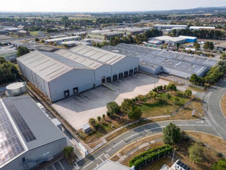 Aerial view of a large industrial warehouse with multiple loading bays, surrounded by roads, trees, and other industrial buildings.