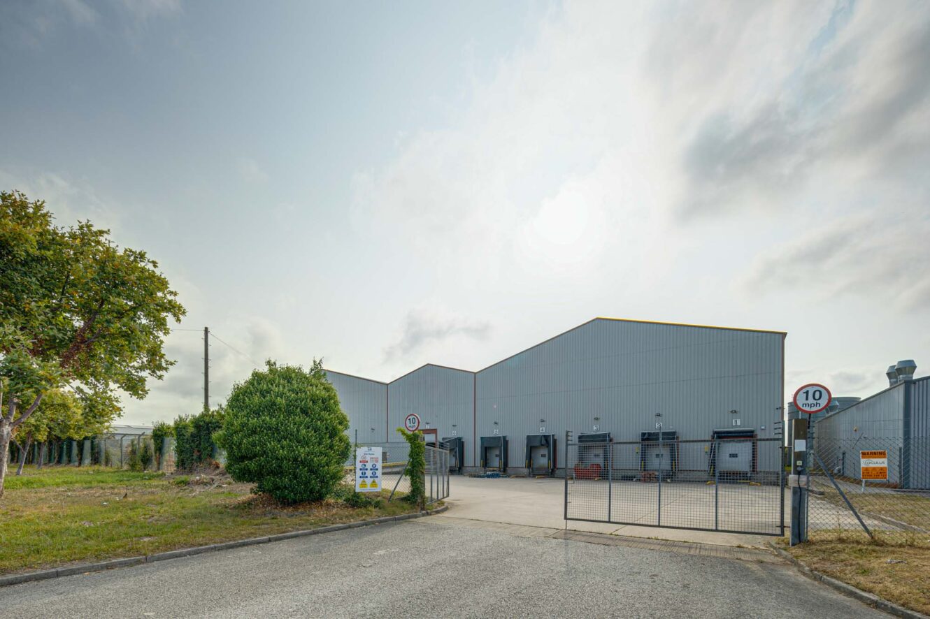 A large industrial warehouse with a gated entrance, multiple loading bays, a speed limit sign, and trees along the perimeter.