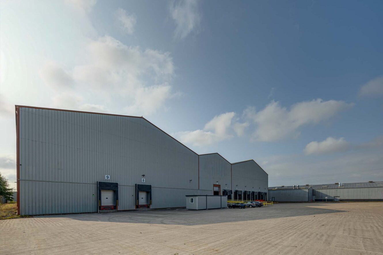 Large industrial warehouse with multiple loading bays and lorries parked outside under a partly cloudy sky.