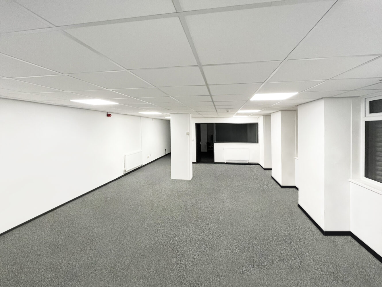 Empty office space with white walls, a suspended ceiling, grey carpet, and fluorescent lighting. There are radiators and windows along the walls.