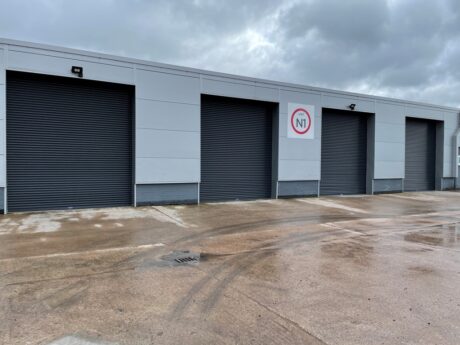 Four closed industrial garage doors on a grey building with an N1 sign above one door; wet concrete ground and cloudy sky.