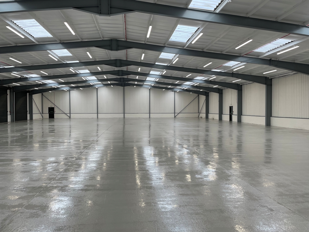 Large, empty warehouse interior with polished concrete floor, metal beams, white panelled walls, and skylights in the ceiling.