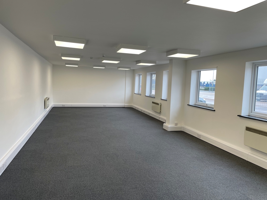Empty office space with grey carpet, white walls, ceiling lights, and multiple windows along one side allowing natural light into the room.
