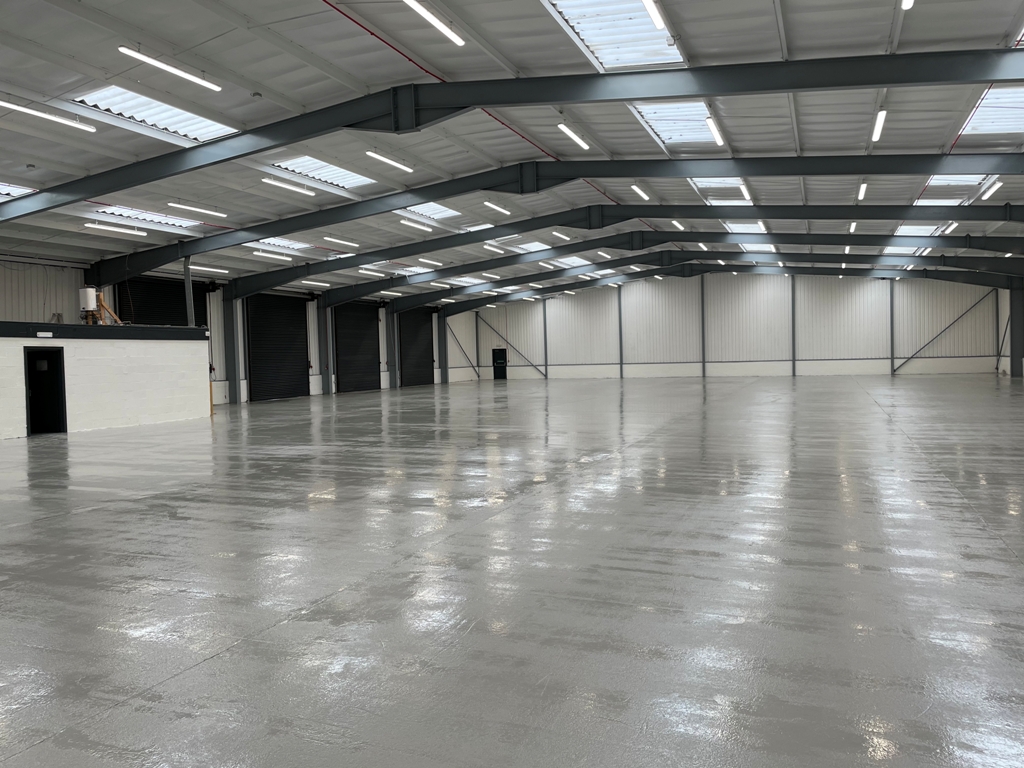 Large, empty industrial warehouse with a shiny concrete floor, high metal ceiling, overhead lights, and a small enclosed office area to the left.