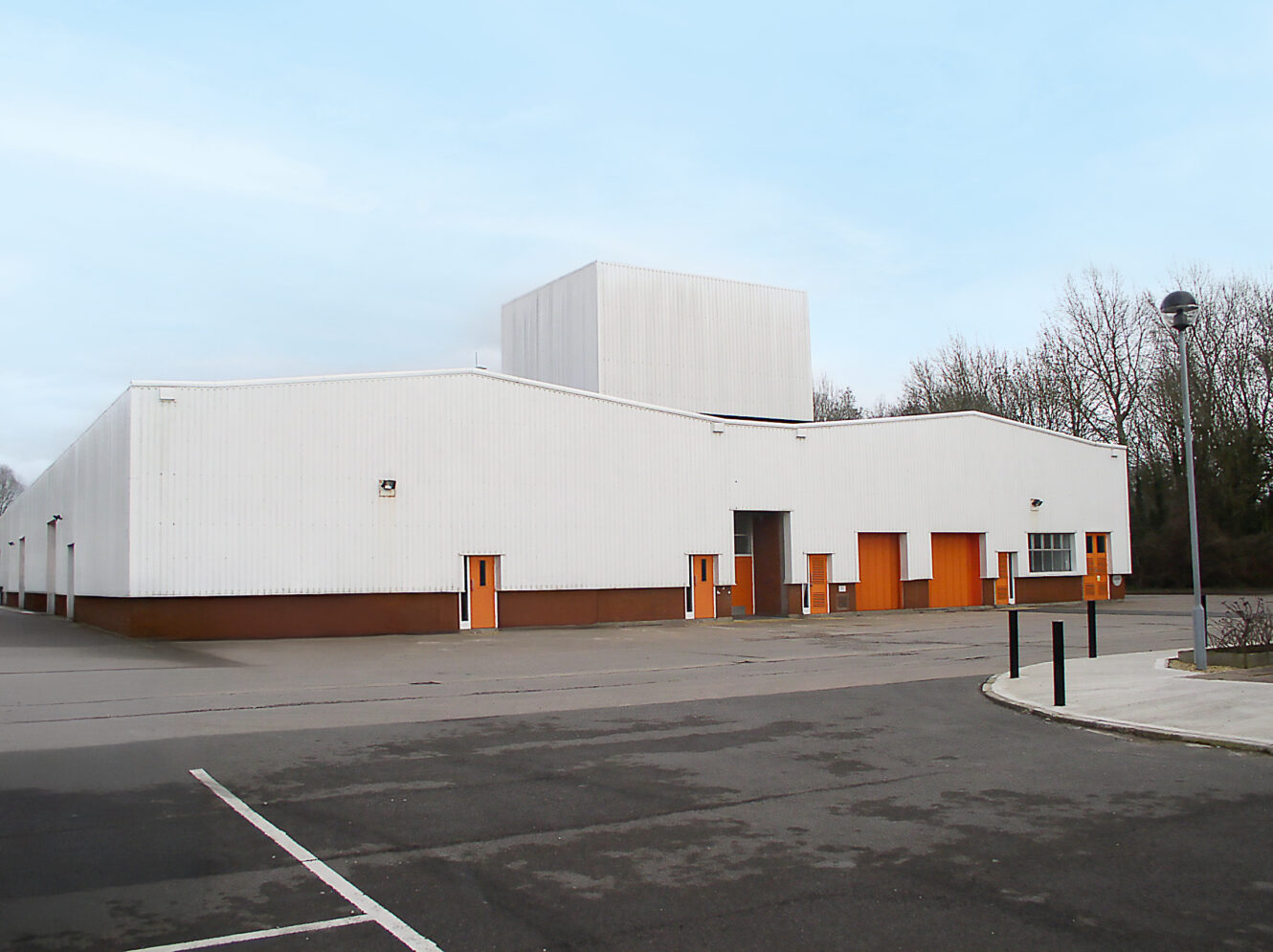 A large, white industrial warehouse with orange doors and minimal windows, surrounded by an empty car park and some trees in the background.