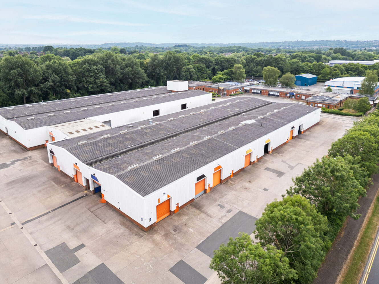 Aerial view of a white industrial warehouse complex with orange doors, surrounded by trees and a large paved area.