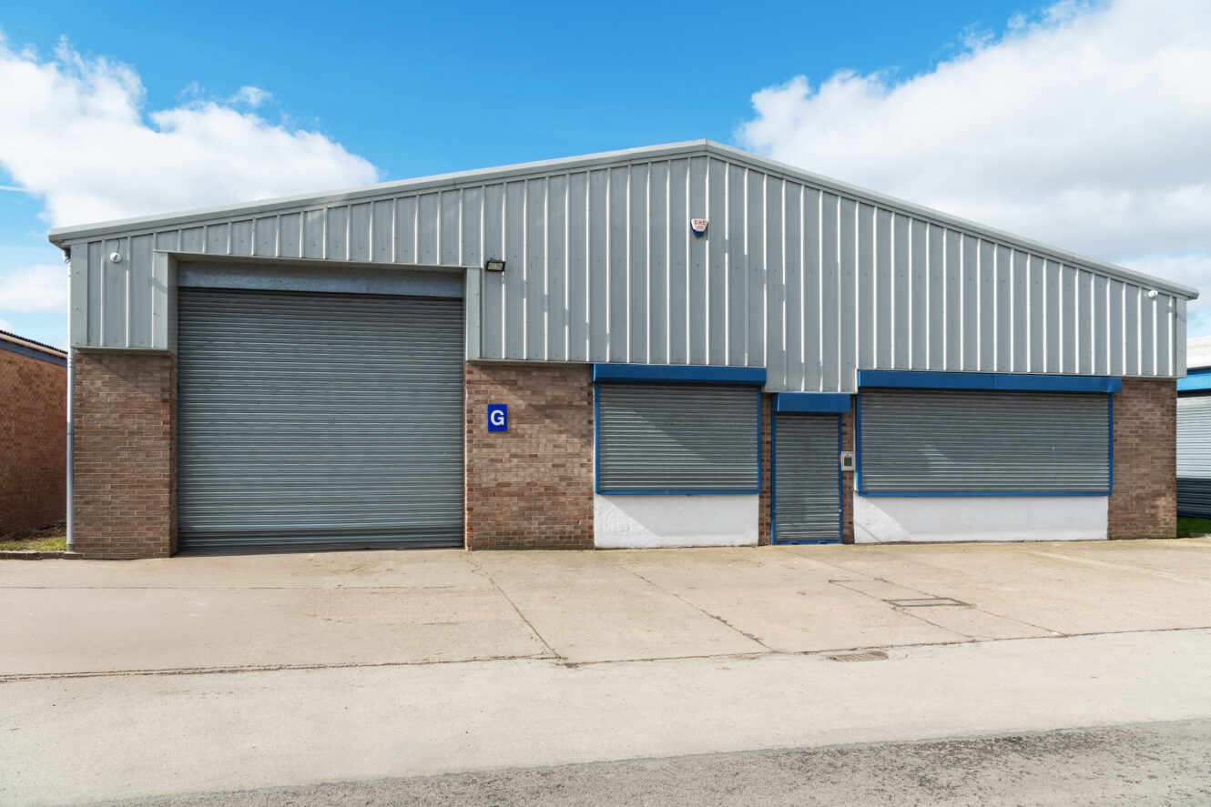 A single-storey industrial warehouse with a large grey roller shutter door, metal cladding, brick lower walls, and a sign labelled G on the front. The sky is partly cloudy.