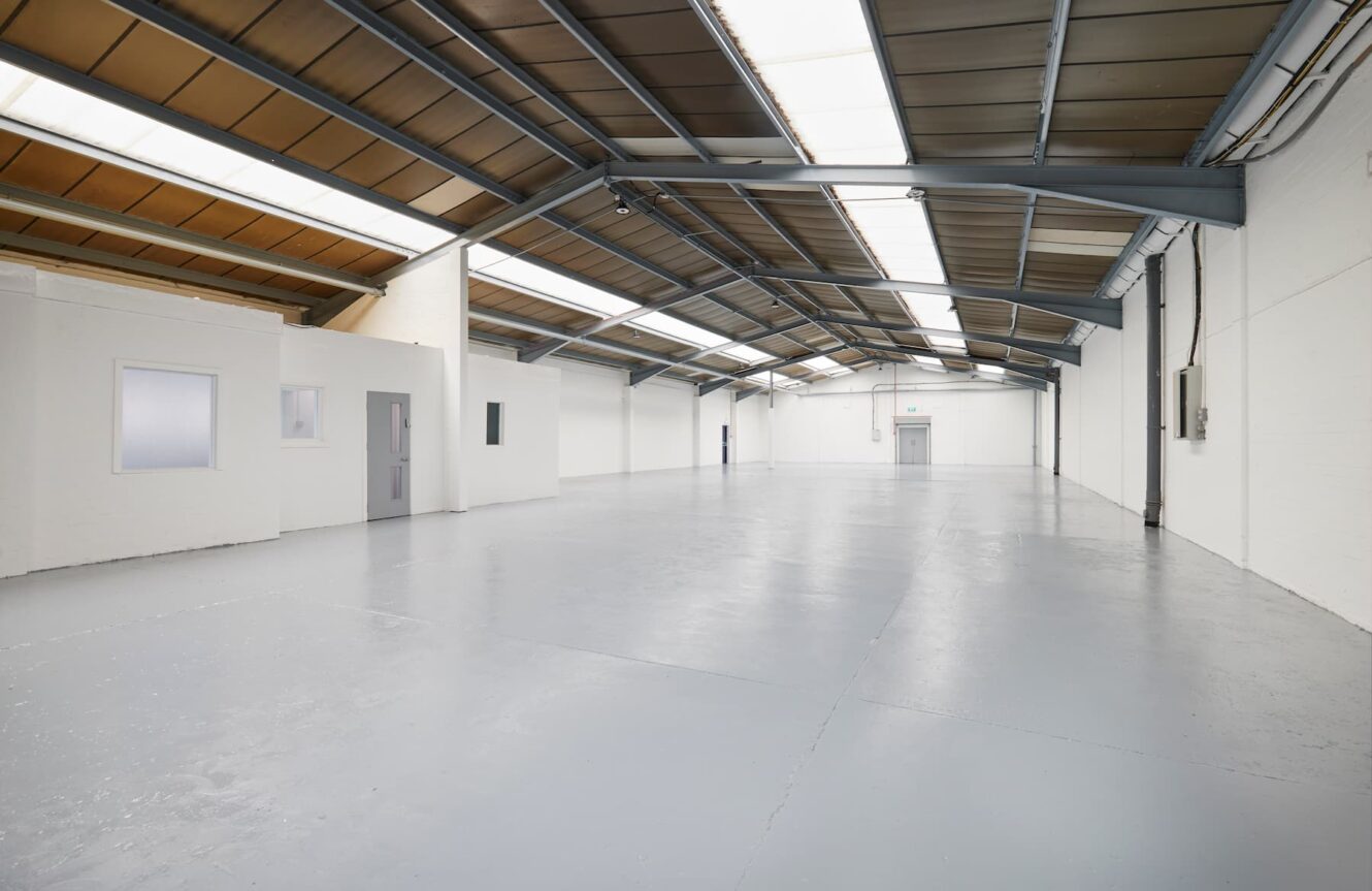 Interior of an empty industrial warehouse with white walls, grey floor, and a high ceiling with rooflights.