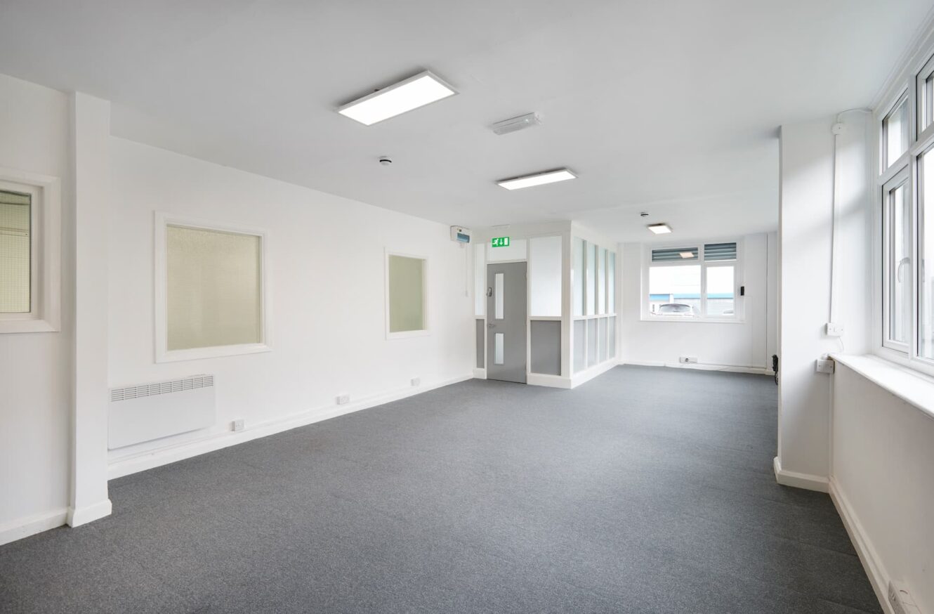 Empty office space with white walls, grey carpet, large windows on the right side, and overhead fluorescent lighting.