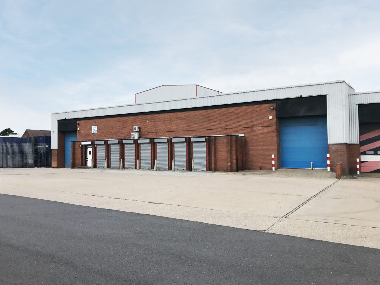 A large industrial warehouse with several closed metal loading bay doors and a blue entrance door, situated on a concrete yard.