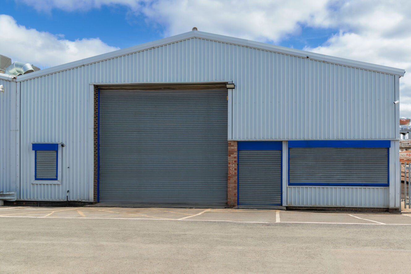 A large industrial warehouse with blue and grey metal exterior, featuring a wide roller shutter door and two small windows, situated in an empty car park.