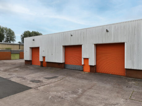 Industrial building with three orange roller shutter doors on a concrete yard, metal cladding, and a clear sky in the background.