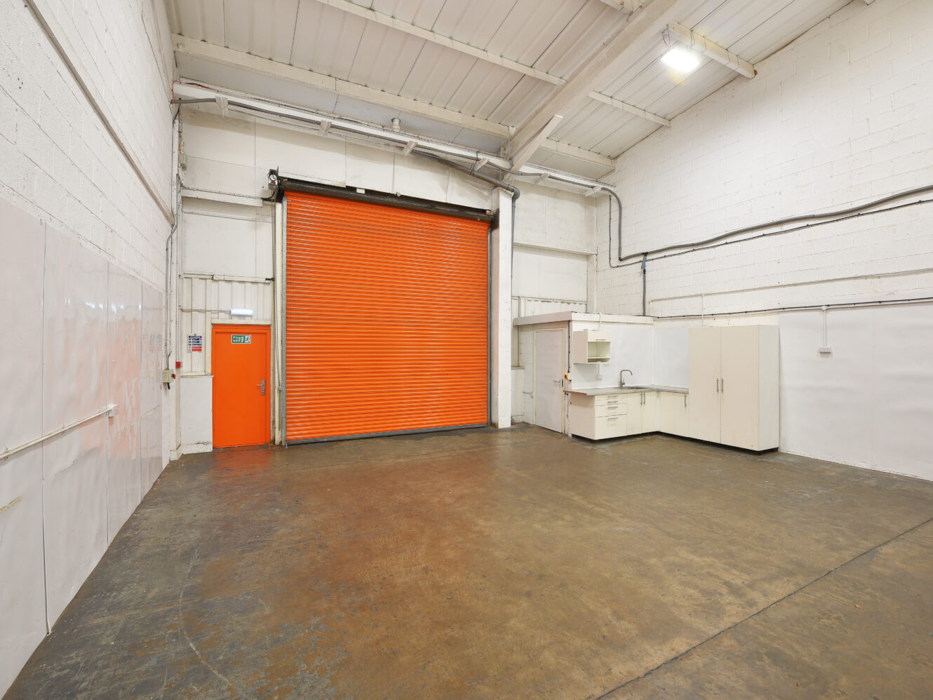 Empty warehouse interior with a large orange roller shutter, a small orange side door, white cupboards, and a concrete floor under bright fluorescent lighting.