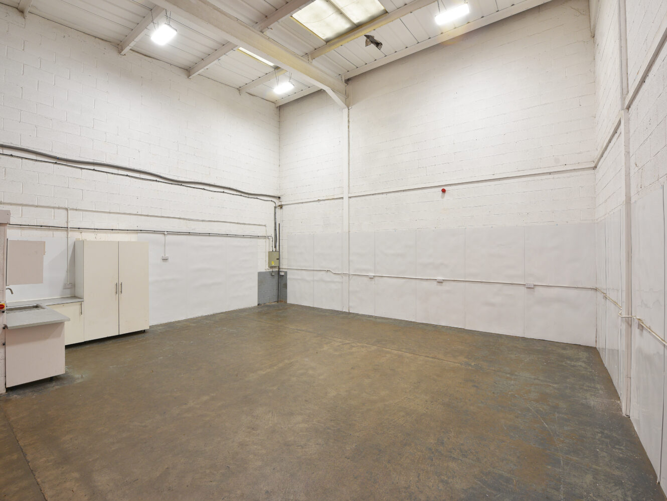 Empty industrial warehouse with white walls, high ceiling, overhead lighting, and a few cupboards and workstations along one wall.